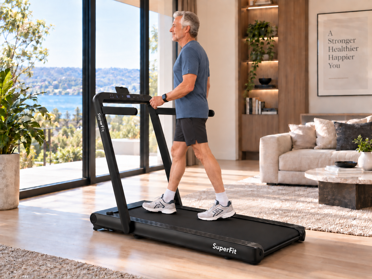 A man walks on a black SuperFit treadmill in a bright, modern living room with large windows overlooking a scenic lake.